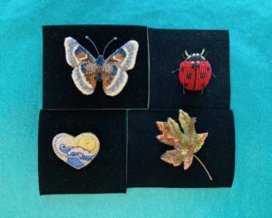 four beaded brooch pins - top left: a butterfly, top right: a ladybug, bottom right: a maple leaf, bottom left: a heart with an image of the ocean horizon