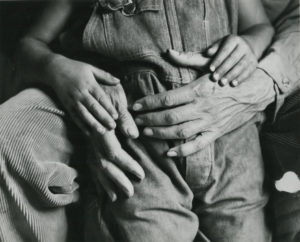 An image of the body of a child in overalls with their hands on the hands of their parent around their waist.