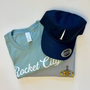 a navy hat with a white Saturn V stitched on, on top of a light teal t-shirt with the words Rocket City in white script, on top of a light blue t-shirt with a cartoon of Eggbeater Jesus.