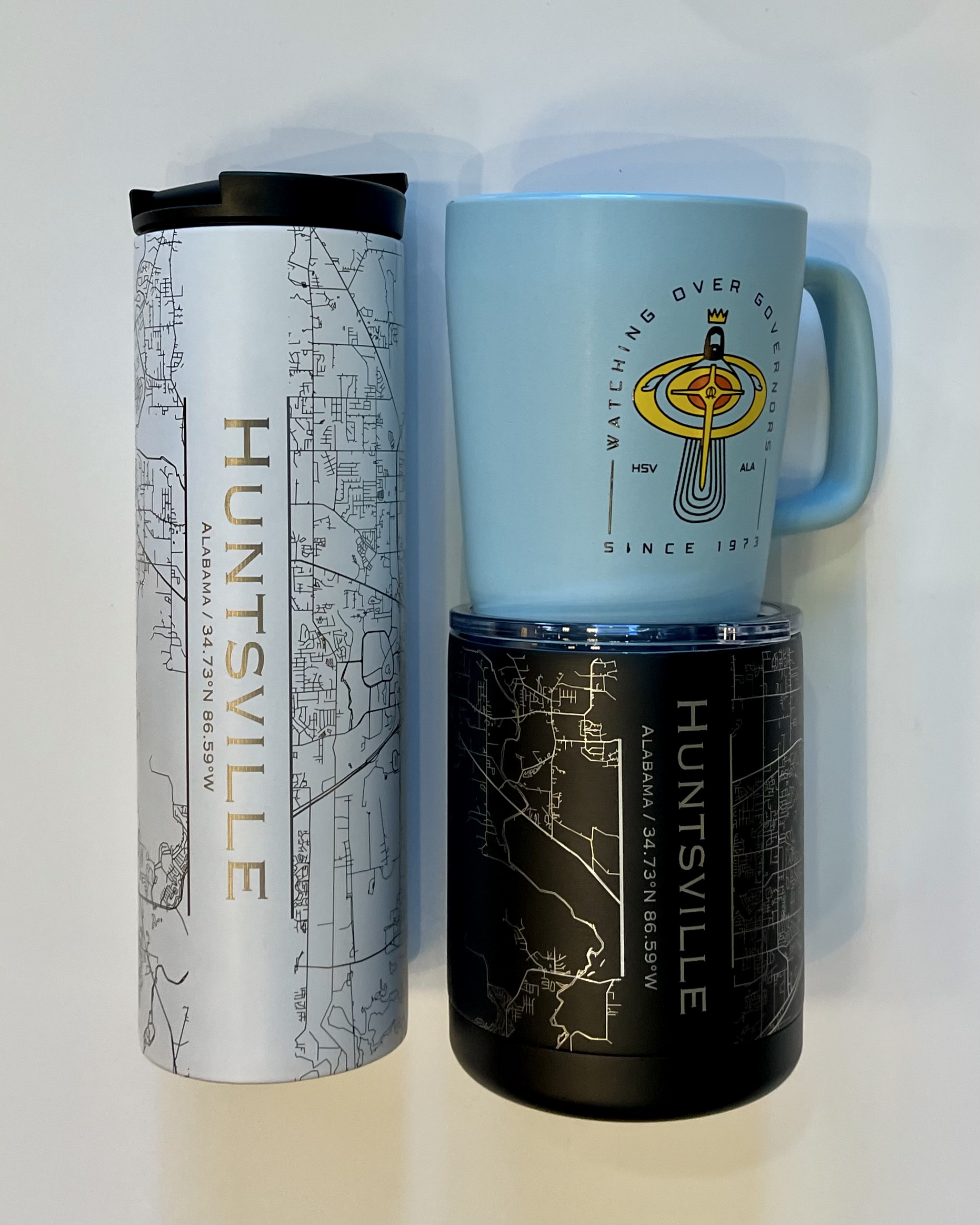 left: tall white insulated bottle with a gold map of Huntsville. top right: light blue mug with the Eggbeater Jesus picture. bottom right: short black tumbler with a silver map of huntsville