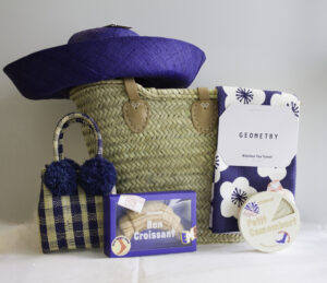 a collection of items for a picnic: a blue straw hat, a big straw bag, a small gingham straw bag, a floral tea towel, and two pairs of Eat My Socks brand socks in a croissant shape and a camembert cheese shape