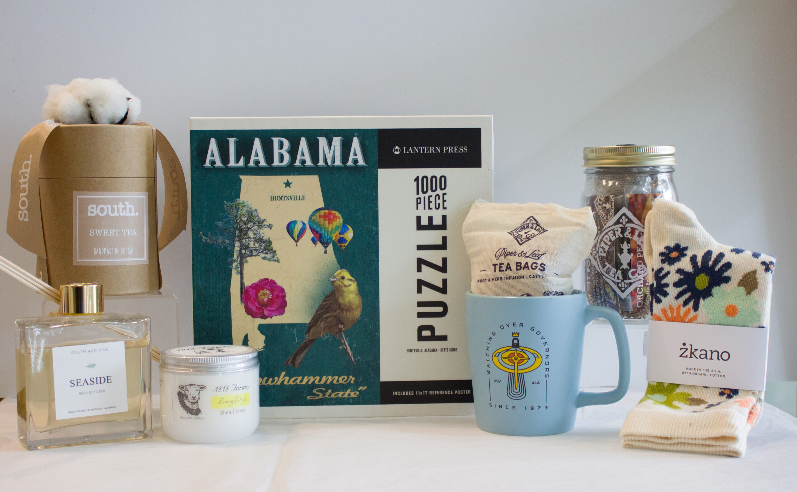 a collection of items from local businesses: candles from South candles, a puzzle in the state of Alabama, tea from Piper & Leaf, a mug from Rocket City Apparel, socks from zkano, skincare from 1818 farms, and a diffuser from South and Pine