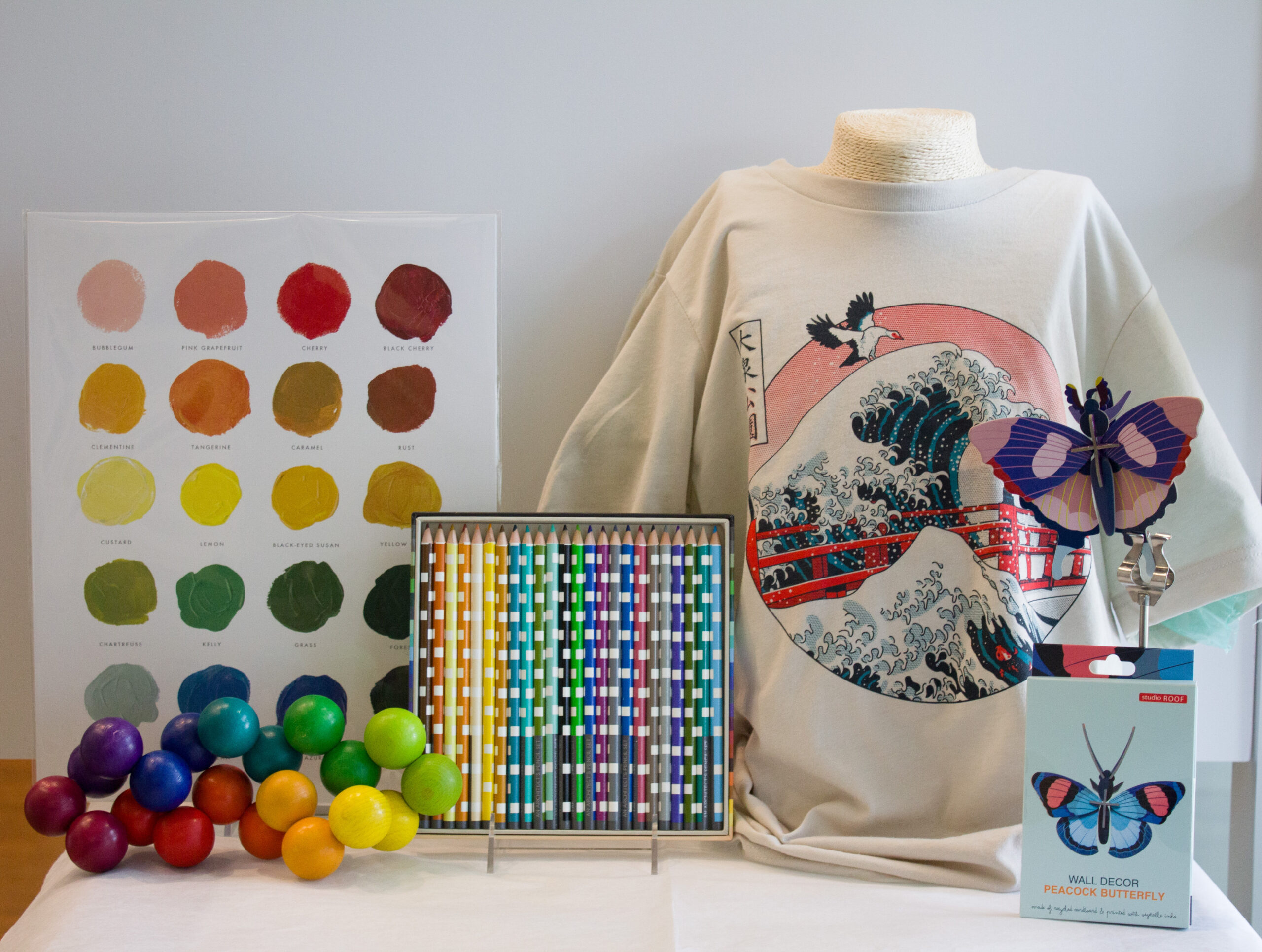 a collection of art-inspired items: a color swatch art print, a t-shirt inspired by The Great Wave off Kanagawa and Big Spring Park, a build-your-own cardboard 3D butterfly, some colored pencils, and a colorful set of wooden spheres